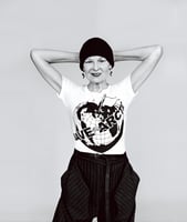 A black and white portrait shows Dame Vivienne Westwood with a toothy grin, c. 2014. The designer is  has elbows out and arms behind her head and is wearing a beanie, earrings, a t-shirt that reads save the artic in bold font over a stylized image of the globe shaped like a heart with a white flag planted in the arctic zone, and stylized pinstripe trousers.