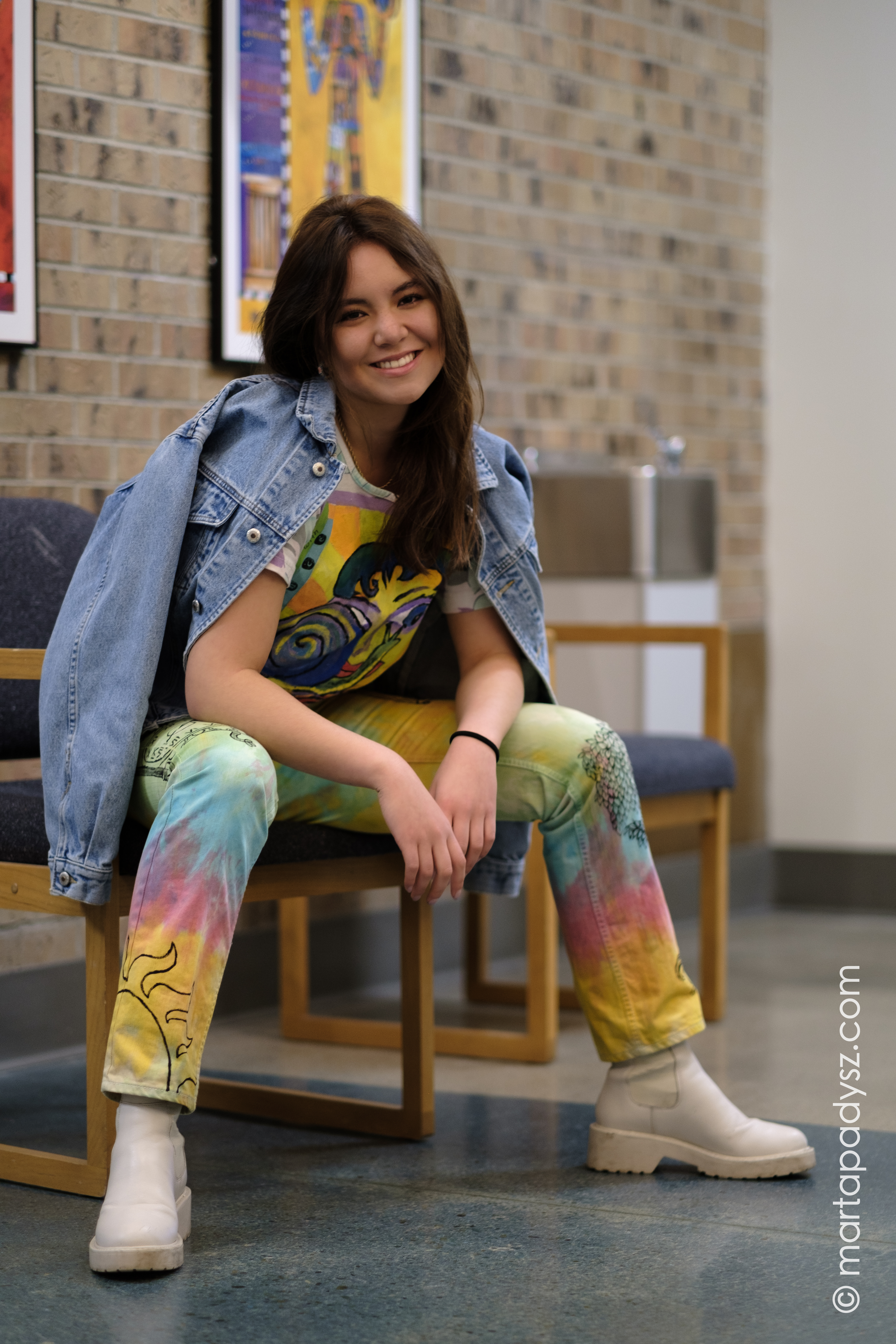 Seated model smiles into the camera, she is wearing colorful tie dye pants and a graphic tee with a denim jacket draped over her shoulders.