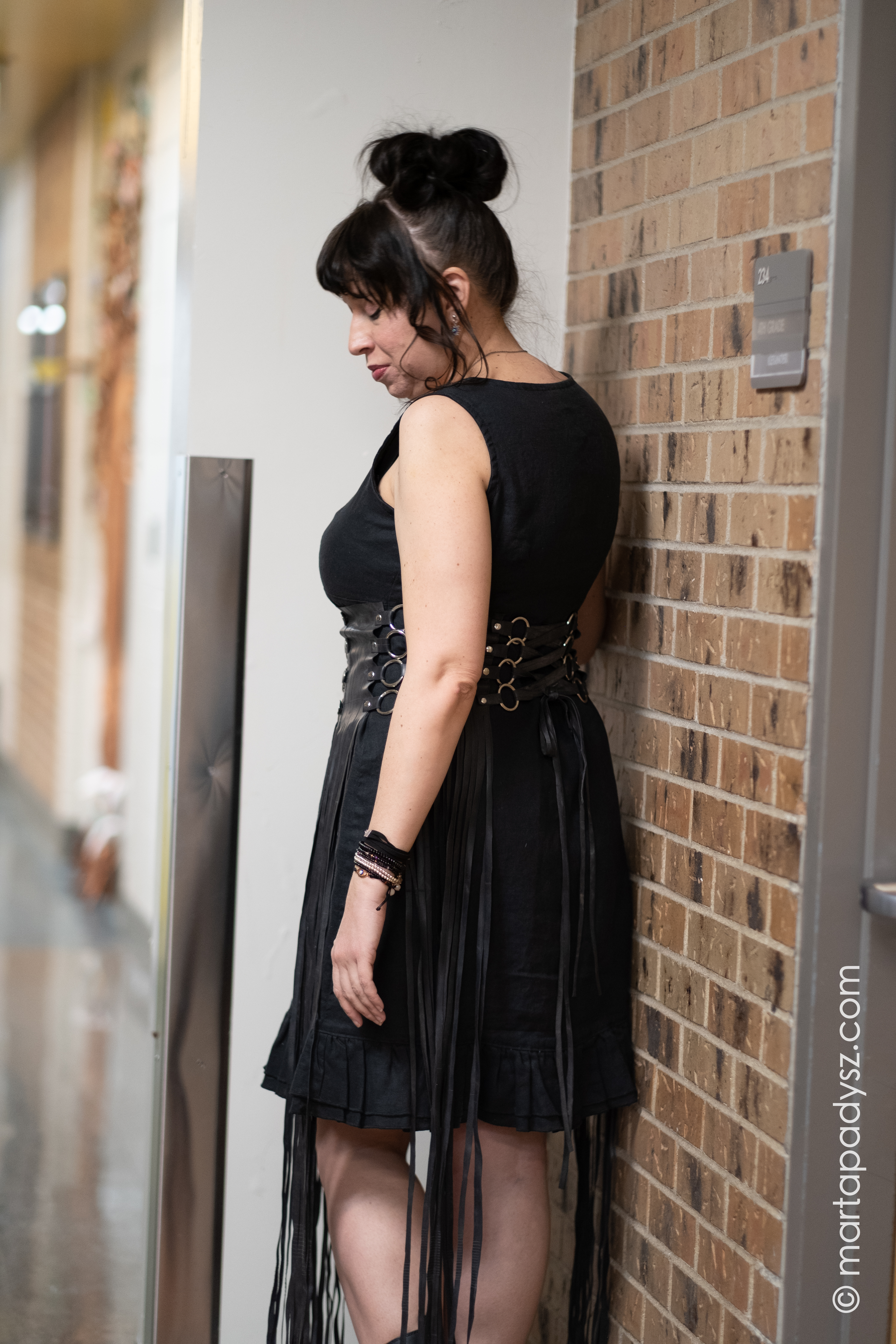 Model Jewel Cavazos is turned 3/4 view with her back to the camera looking over her shoulder towards the ground. She is wearing a black linen sleeveless shift dress from Goddess Gear, accentuated by a stylized corset with nearly floor-length fringe made of upcycled bike tubing by A Subtle Notion.